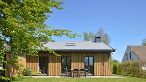 Une maison en bois avec un toit en tuiles, une porte d’entrée et une terrasse avec une table et des chaises.