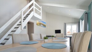 Un espace repas moderne avec une table en bois, des sets de table bleus et des chaises de couleur beige. Un escalier avec des rampes blanches mène à un niveau supérieur.