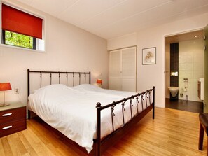 Een slaapkamer met een houten vloer, een metalen bed, een nachtkastje met een lamp, een raam met een rode rolgordijn, een deur naar een badkamer en een lijst met een afbeelding aan de muur.