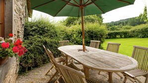 A wooden outdoor table with a green umbrella, surrounded by wooden chairs, set against a stone wall with a garden view. - Rustic Vacation Home, Baneux (Lierneux)
