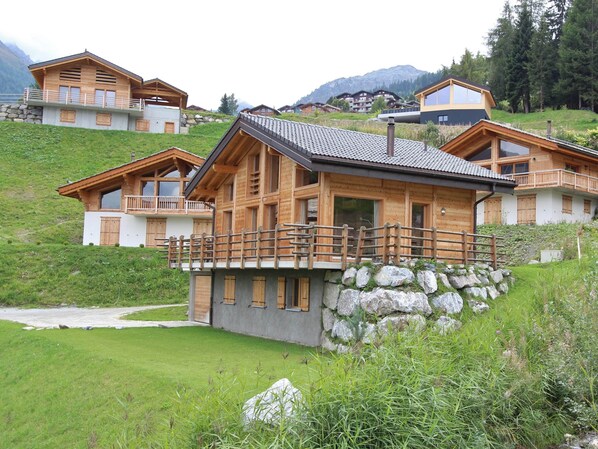 A wooden chalet with a balcony, set on a hillside with a view of other chalets and mountains.