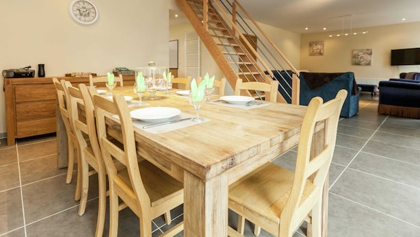 Comedor con una mesa de madera dispuesta para seis personas, rodeada de sillas de madera. Una escalera conduce a un nivel superior.