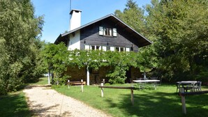 Une maison rustique avec des fondations en pierre, un toit en bois et une cheminée. Un chemin de gravier mène à la maison, qui est entourée d’arbres et agrémentée d’un banc.