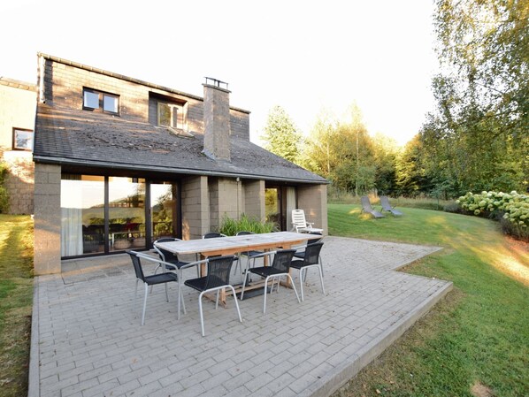 A modern house with a patio area featuring a wooden table and chairs, surrounded by a well-maintained lawn and trees.