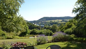 A wooden table with four chairs in a garden setting with a view of a church and hills in the background. - Cozy Villa in Vielsalm With Private Garden (Vielsalm)