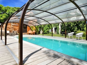 Une piscine couverte dotée d’une terrasse en bois, de chaises longues et d’une structure fermée par des parois de verre.