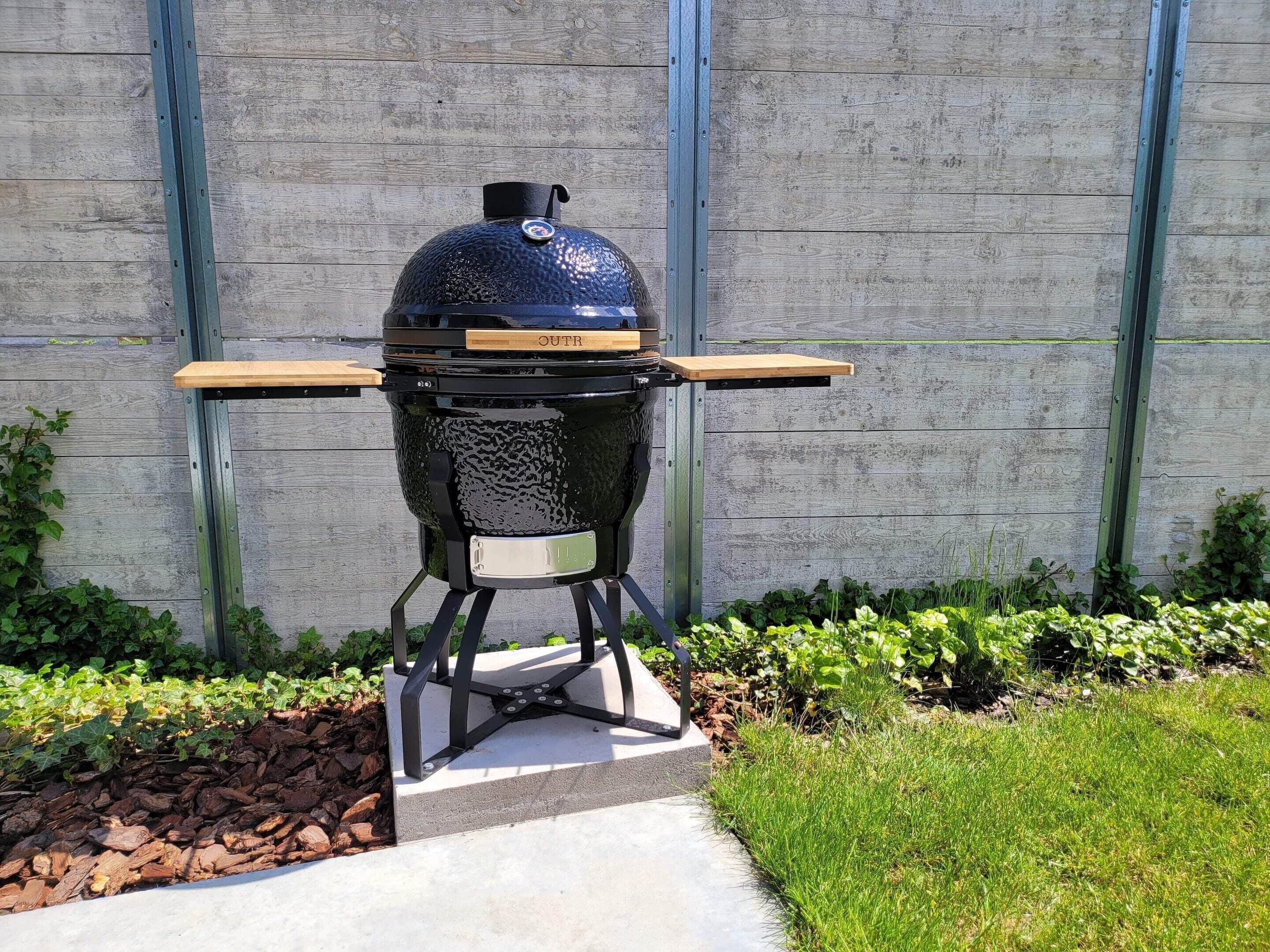 A portable barbecue grill with a lid, two side shelves, and a control panel, set against a wooden fence.