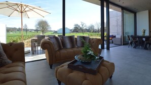 A spacious living area with a large glass door leading to an outdoor patio. Inside, there is a brown sofa with cushions, a coffee table with a glass bowl and flowers, and a wicker chair. - Modern Villa Stay in Dalhem (Dalhem)
