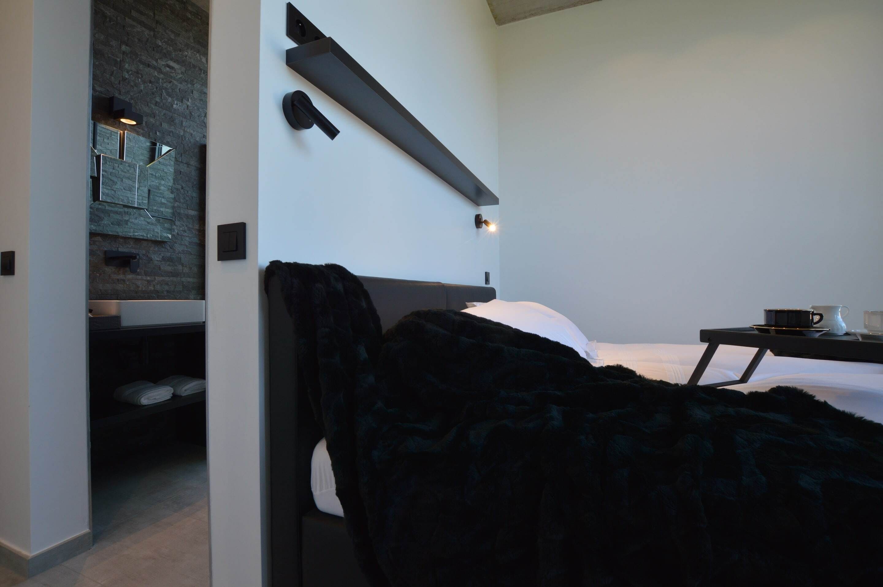 A modern hotel room with a bed, a bedside table with a cup and a book, a wall-mounted shelf, and a dark-colored blanket.