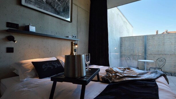 A modern hotel room with a bed, a bedside table with a champagne bottle, a glass, and a blanket. There is a large window with a view of a building and a concrete wall. - Modern Villa Stay in Dalhem (Dalhem)