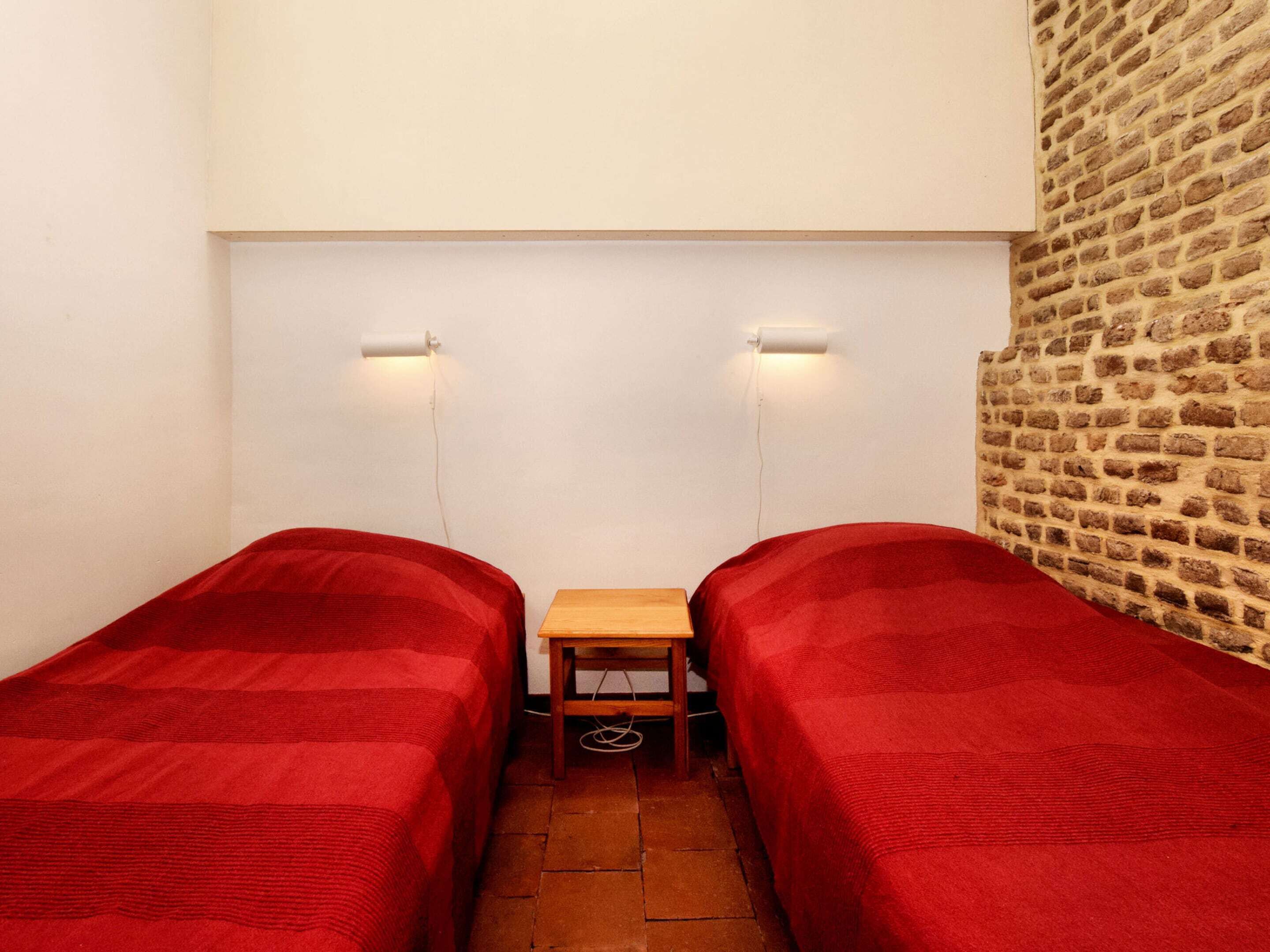A small, narrow room with two single beds, each with a red blanket. There is a small wooden table between the beds, and a brick wall on one side.