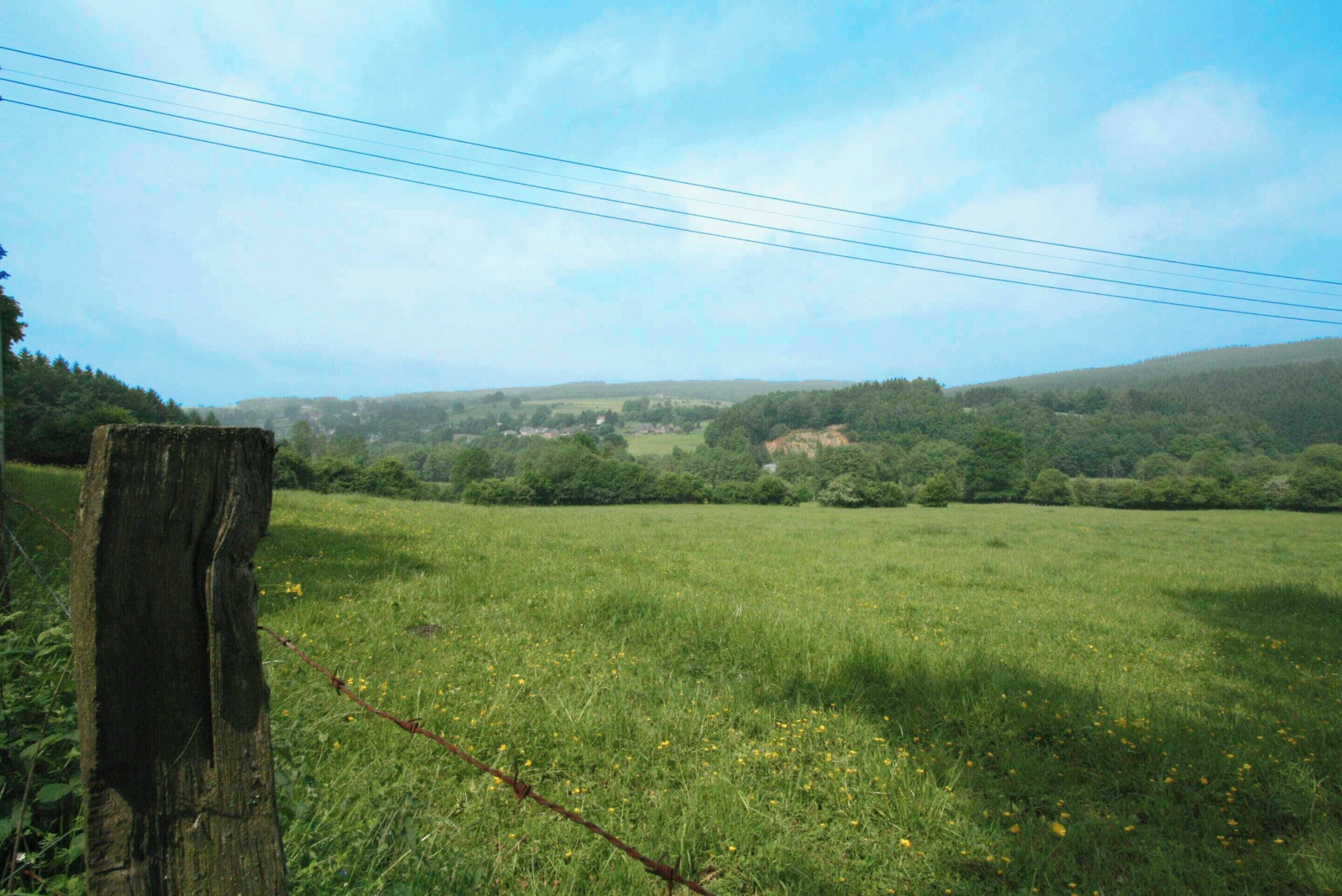 A rural landscape with a wooden fence, a grassy field, and a distant view of hills and a small settlement.