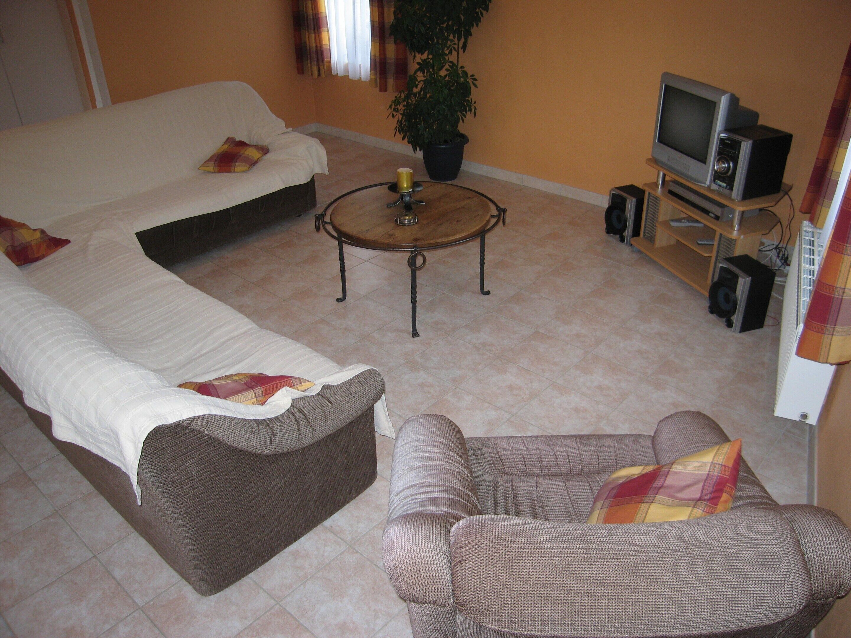 A living room with a sofa, armchair, coffee table, TV, speakers, and a plant.