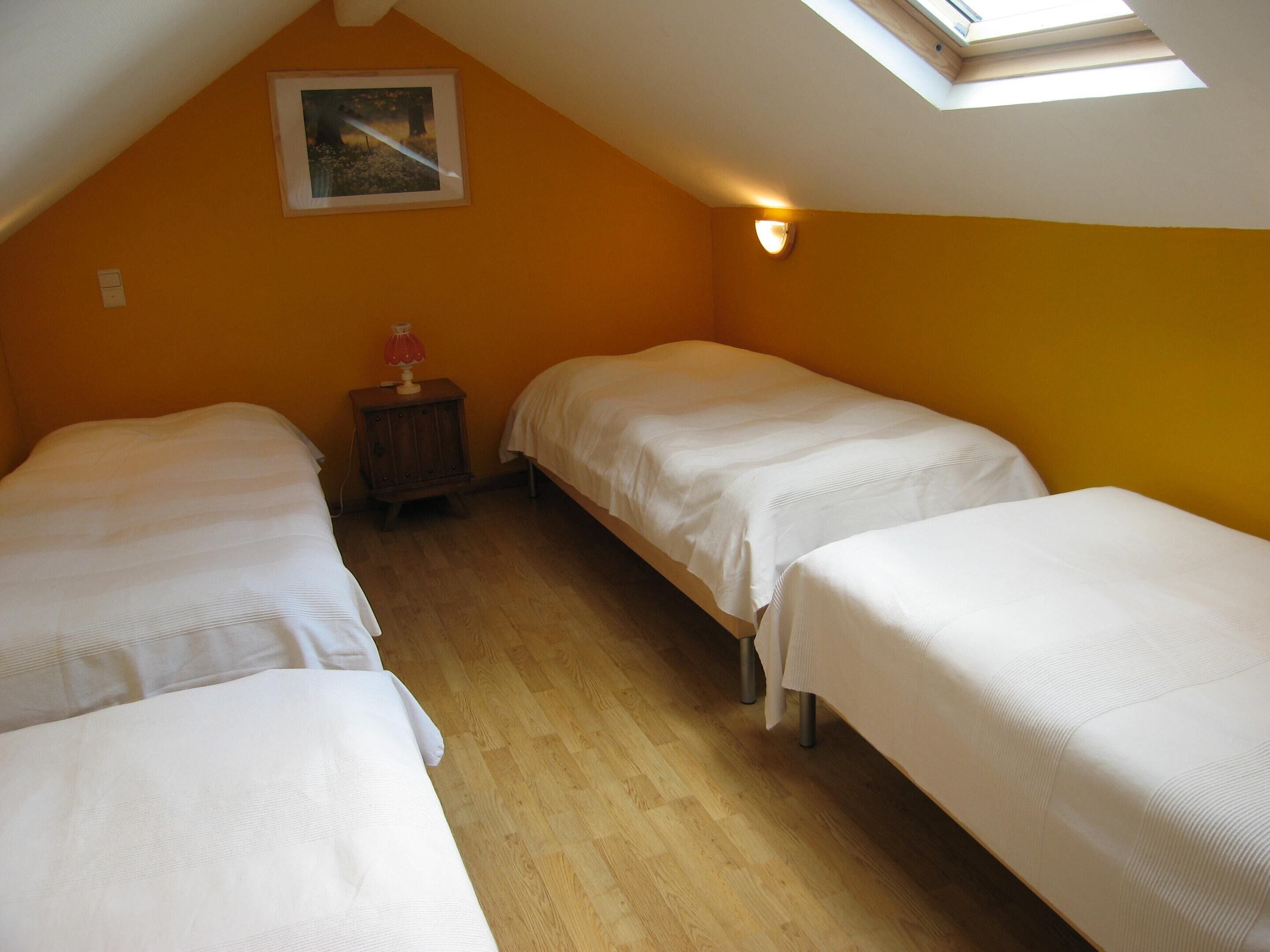 Une chambre avec trois lits, un plancher en bois, une petite table avec une lampe et un puits de lumière.