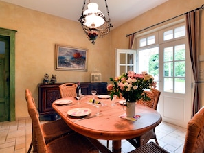 A dining area with a round wooden table set for four, surrounded by wicker chairs. There is a painting on the wall, a chandelier above the table, and a window with curtains to the right.