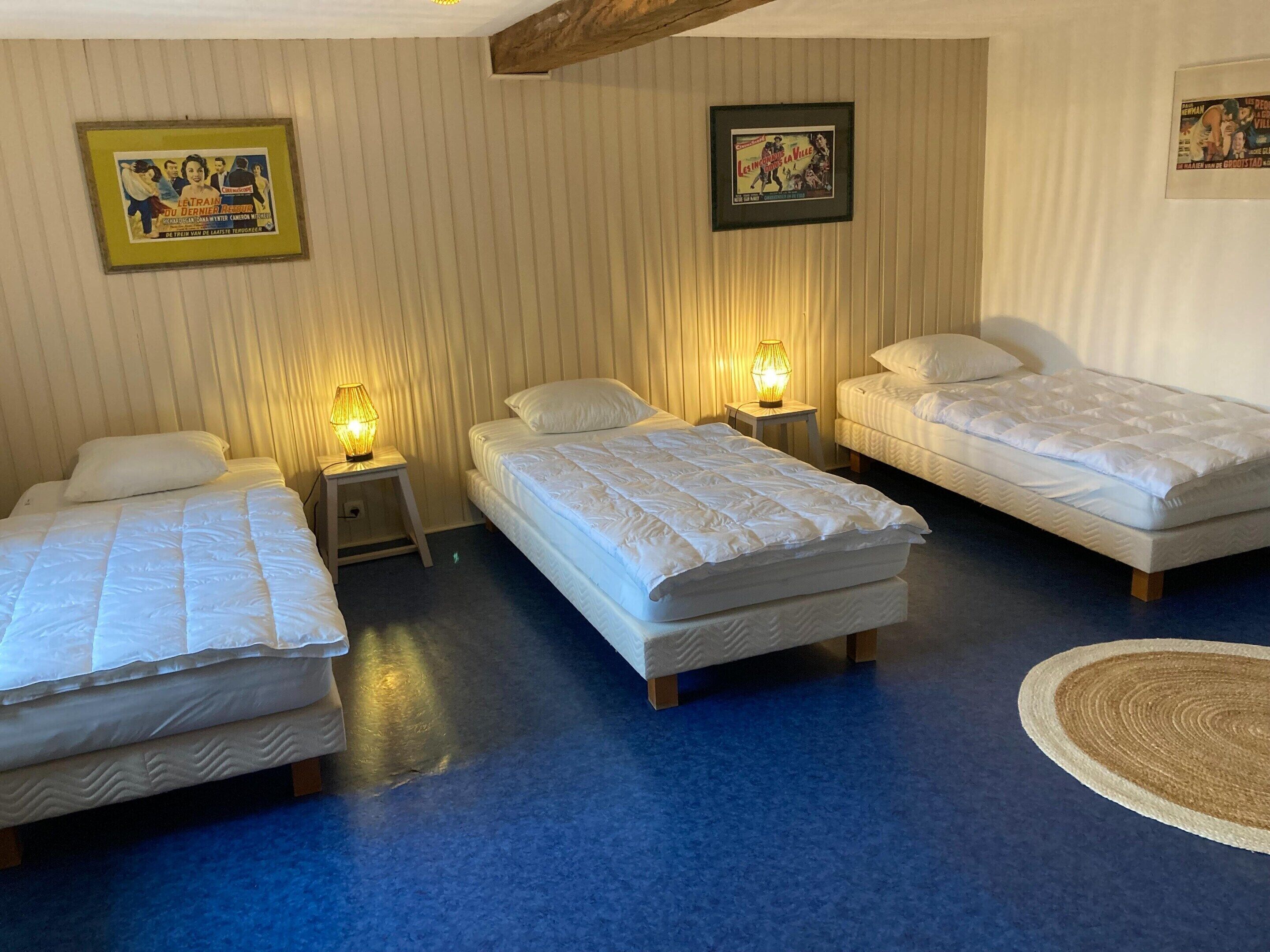 A hotel room with three beds, a small table with a lamp, and two framed pictures on the wall.