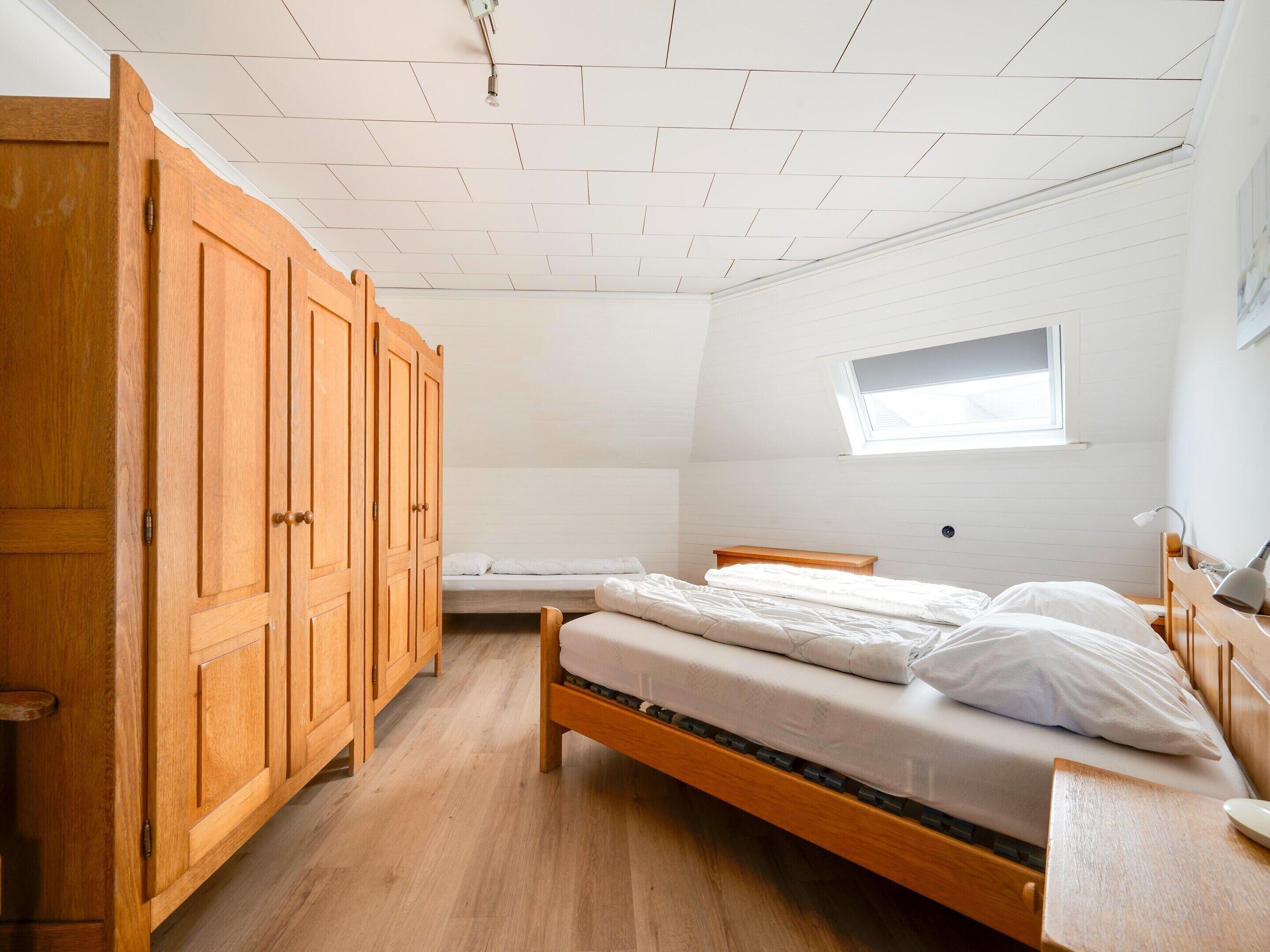 Une chambre à coucher avec une armoire en bois, un lit simple, une petite fenêtre et un bureau avec une lampe.
