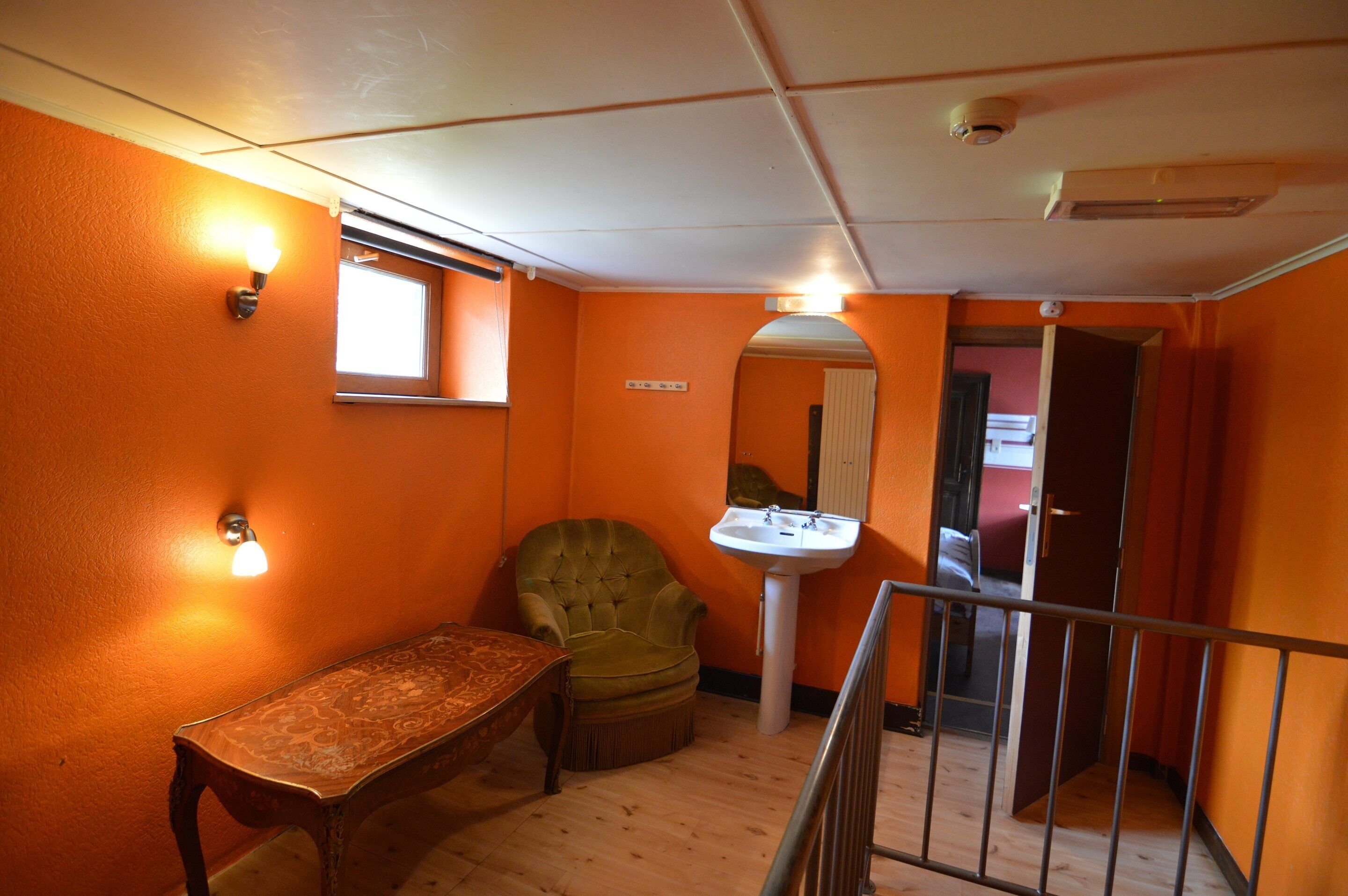A room with orange walls, a wooden bench, a green armchair, a white sink, and a window with blinds.