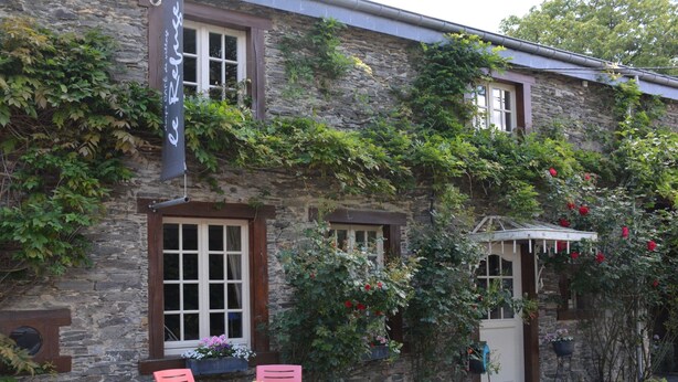 A stone building with a garden area featuring a table and chairs, a hanging sign, and a small covered porch with red flowers. - Holiday Home in Bievre Near Town Center (Bievre)