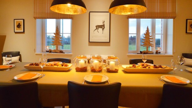 A dining room with a large table set for a meal, two pendant lights, and windows with views of trees. - Charming Fens Family Home (Waimes)