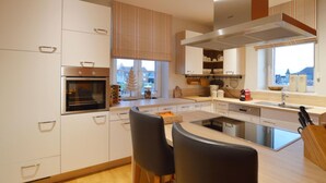 A modern kitchen with white cabinets, a built-in oven, a large island with a sink, and a view of the city through the window. - Charming Fens Family Home (Waimes)