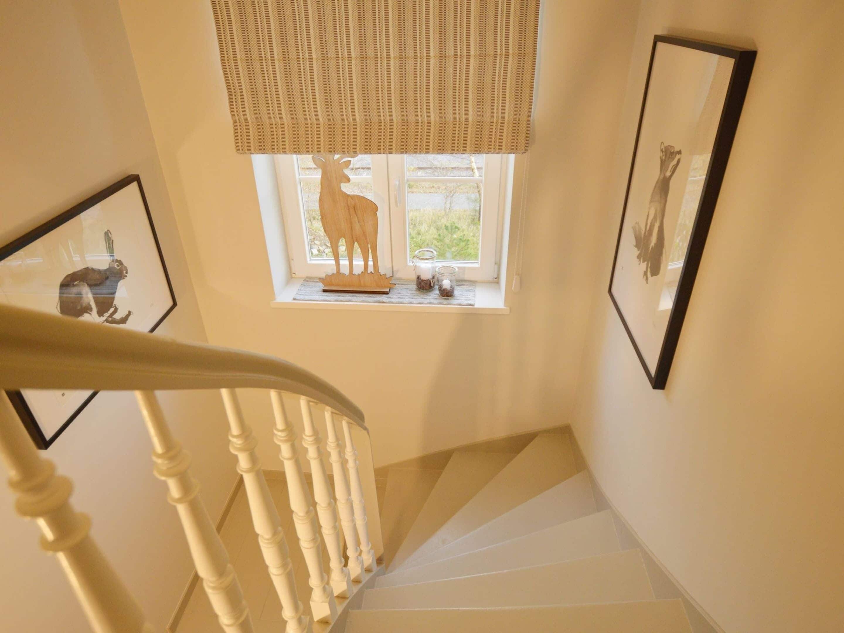 A staircase with a white railing, a window with a wooden deer sculpture, and two framed pictures on the walls.