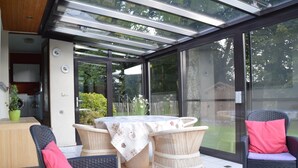 A covered patio area with wicker furniture, a table with a floral tablecloth, and a view of the garden through large glass doors. - Elegant Villa in Stavelot With Fitness and Playroom and an Incredible Garden (Stavelot)