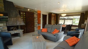 A modern living room with a stone fireplace, grey sofas with orange cushions, and a dining area with a table and chairs. - Elegant Villa in Stavelot With Fitness and Playroom and an Incredible Garden (Stavelot)