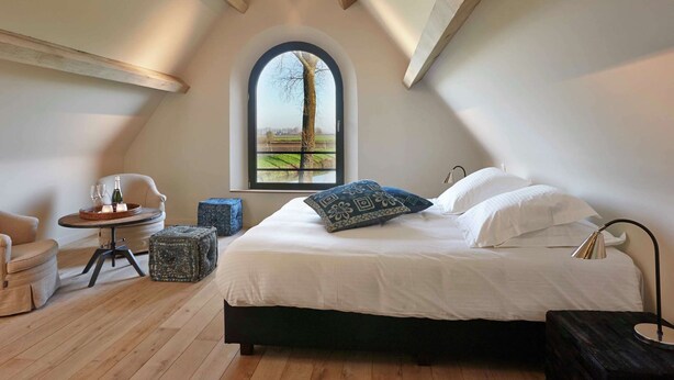 A bedroom with a large bed, a round table with a bottle and glasses, a chair, and a view of a tree through a window. - Country House in Sint Laureins Near Het Zwin (Sint-Laureins)