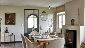 A dining area with a wooden table, wicker chairs, a fireplace, and a large window. - Country House in Sint Laureins Near Het Zwin (Sint-Laureins)