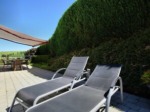 A patio with two lounge chairs and a table, surrounded by a hedge and a view of the countryside.