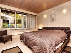 A bedroom with a large bed, a window with blinds, a chair, and a nightstand with a lamp.