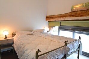 A bed with a metal frame and a white comforter, a bedside lamp, and a window with a view of greenery.