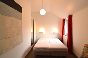 A small, single-bed room with a sloped ceiling, a wall-mounted light fixture, and a window with red curtains.