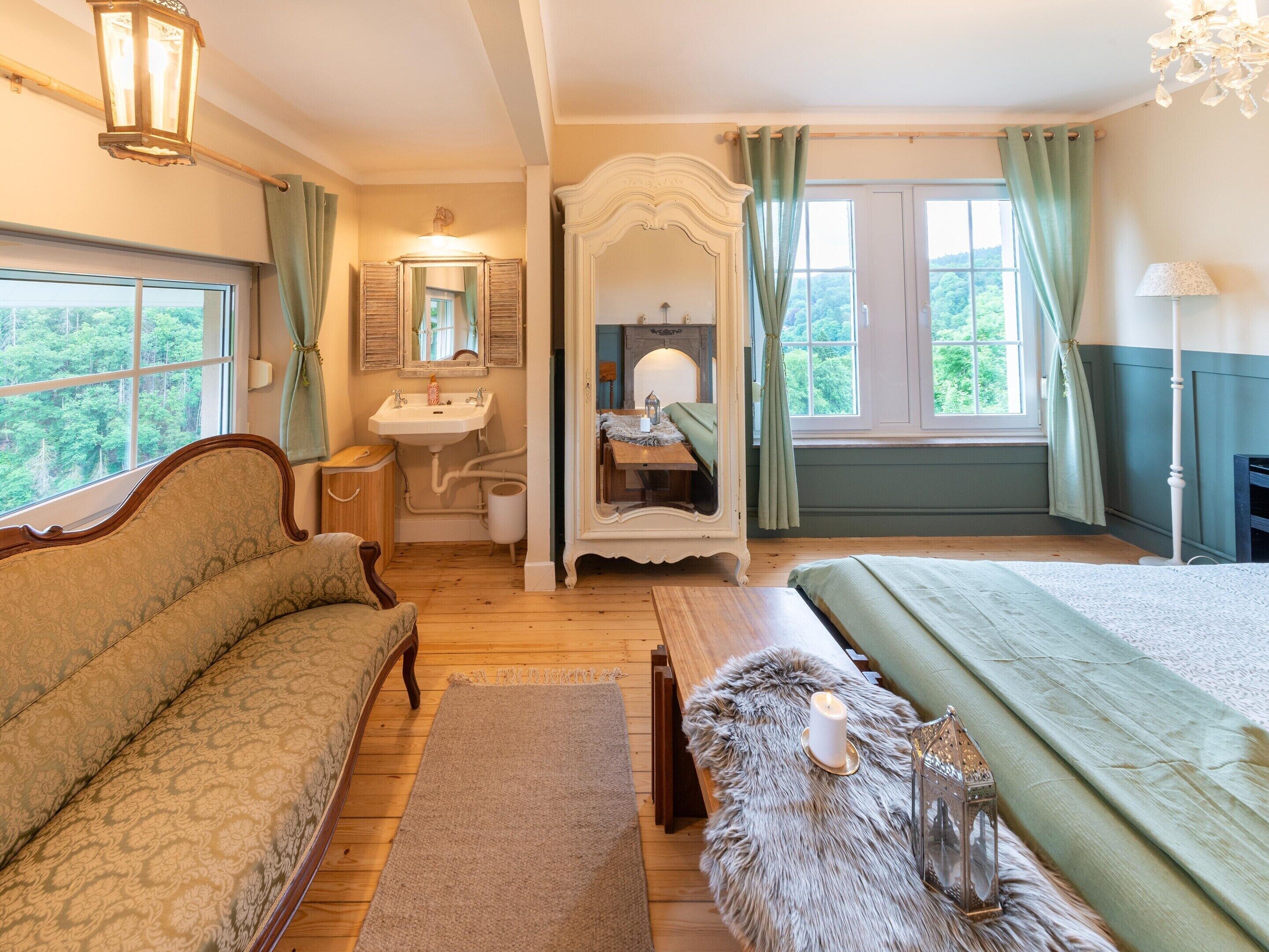 A bedroom with a bed, a sofa, a vanity with a mirror, a sink, and a window with curtains.