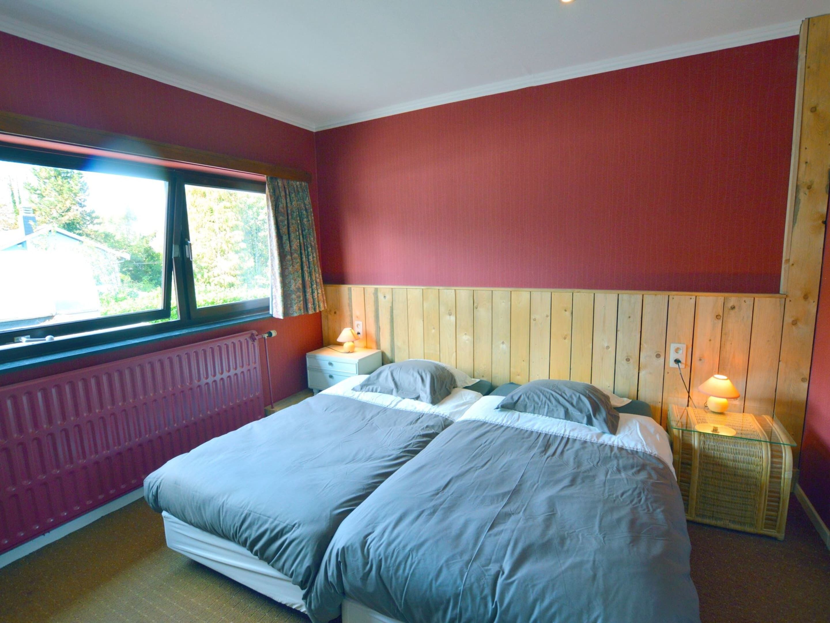 A bedroom with two beds, a wooden headboard, a window with curtains, a bedside table with a lamp, and a radiator.