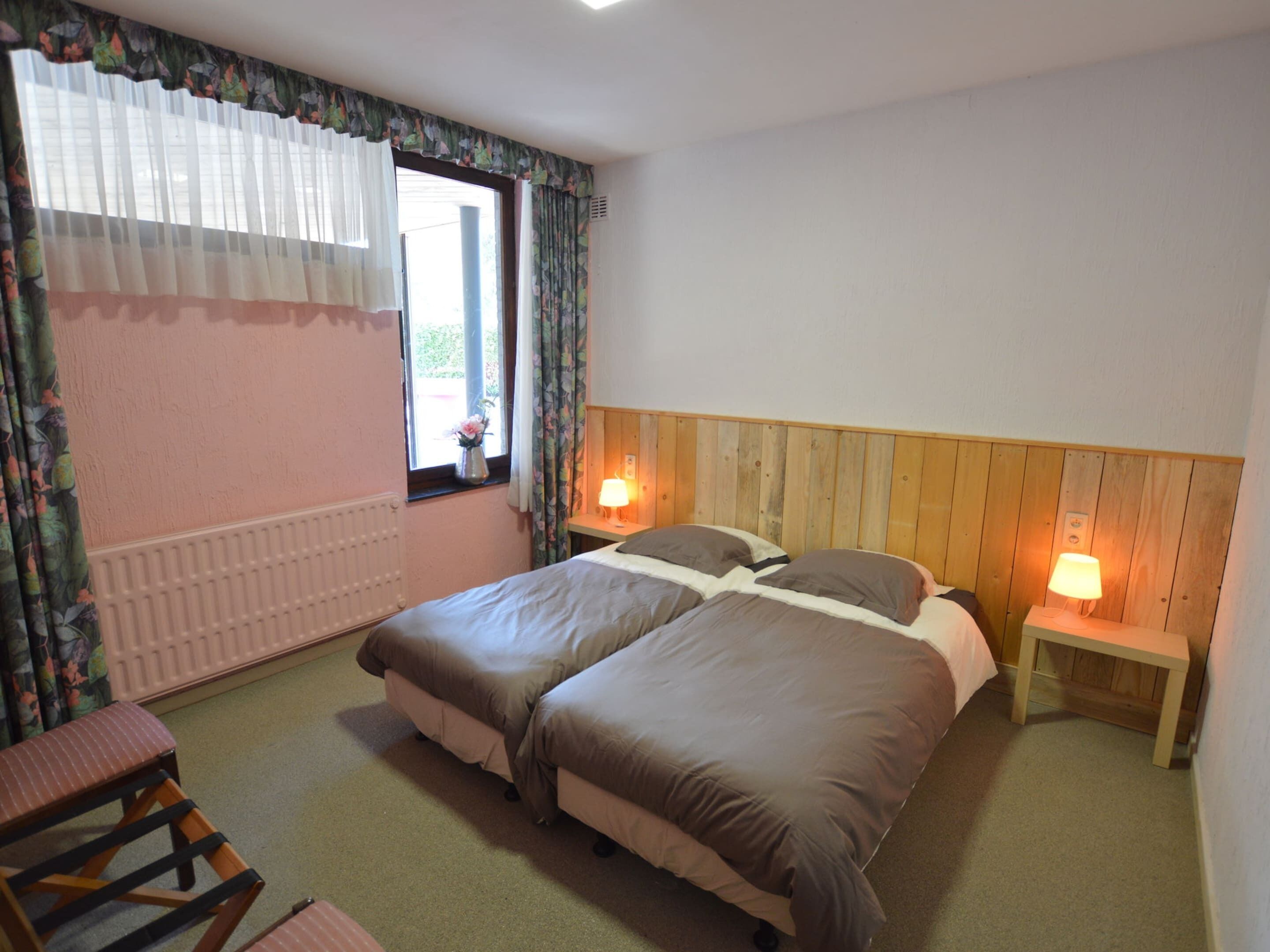 A bedroom with two beds, a wooden headboard, bedside lamps, a radiator, a window with curtains, and a small table.