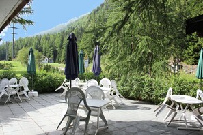 An outdoor patio with white plastic chairs and tables, green umbrellas, and a view of a forested hillside. - Chalet in Valais Near Ski Lift & Saas Valley (Saas-Balen)