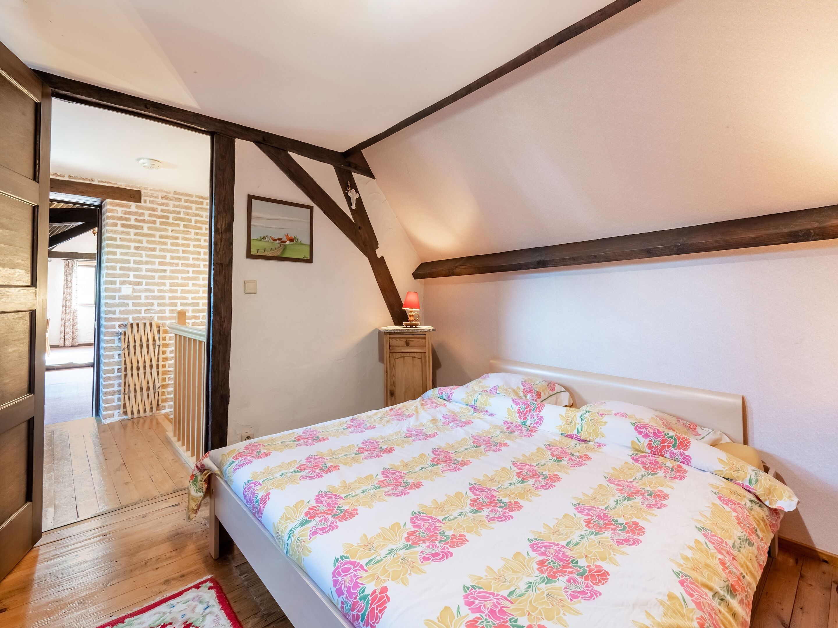 A bedroom with a wooden bed, a nightstand, a picture on the wall, and a view of the outside.
