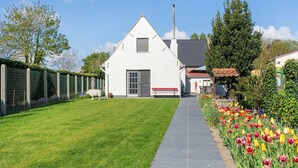 Een wit gebouw met een grijze deur en een raam, een schoorsteen en een pad dat naar een tuin met tulpen leidt.