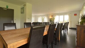 Une salle de conférence avec une longue table en bois et des chaises noires, de grandes fenêtres avec des rideaux et une plante posée sur un meuble en bois.