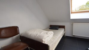 Une petite chambre avec un lit simple, un plafond en pente, une chaise, un radiateur et une fenêtre.