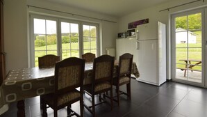Un coin repas avec une table et des chaises, un réfrigérateur et une vue sur un champ verdoyant grâce à de grandes fenêtres.