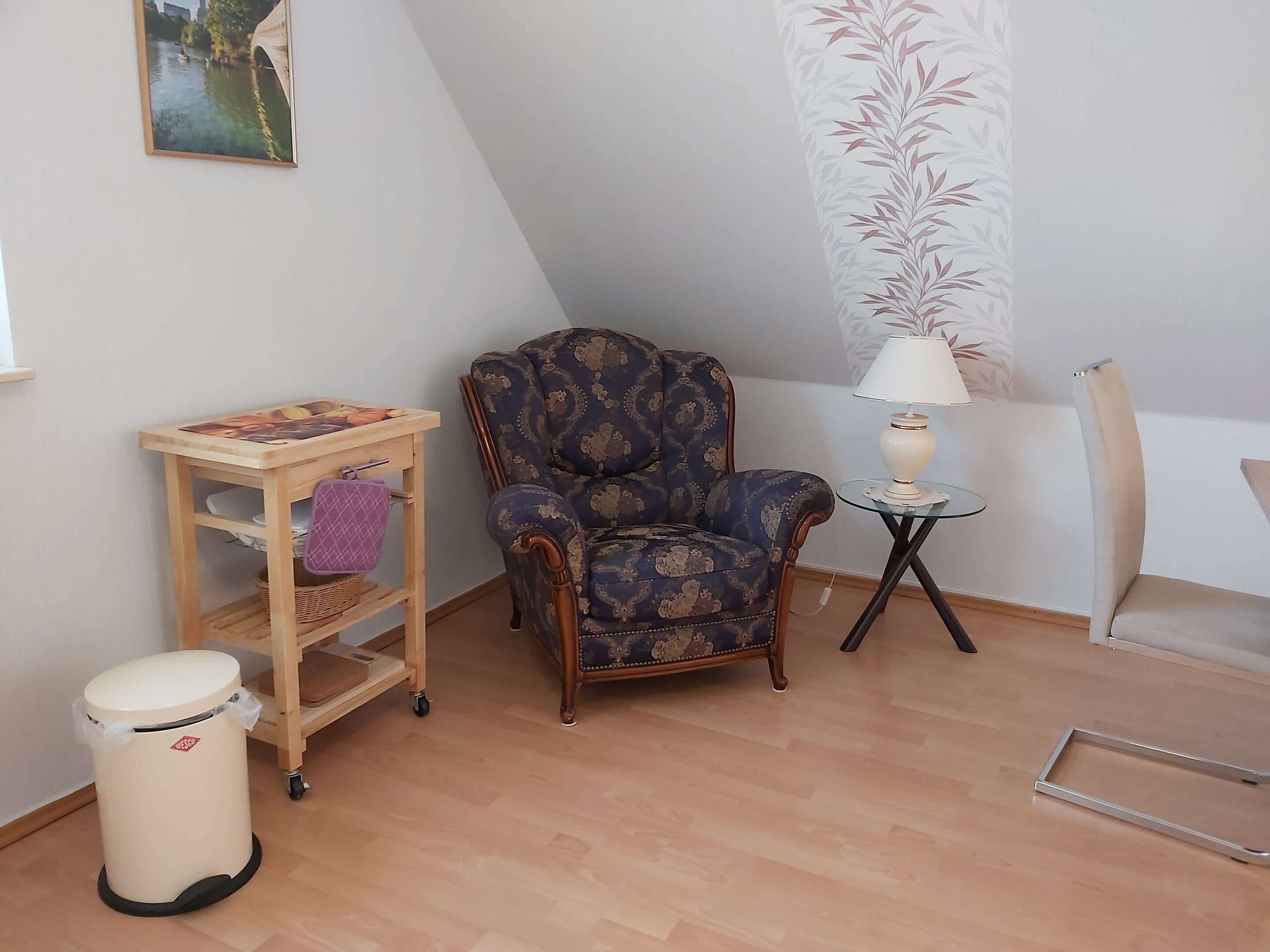 Una habitación con un sillón estampado, una pequeña mesa de madera con una lámpara, un cesto de basura y un cuadro colgado en la pared.