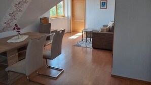 Uma sala de estar moderna com área de jantar, apresentando uma mesa de jantar de madeira, cadeiras brancas, um sofá e uma pequena mesa lateral.