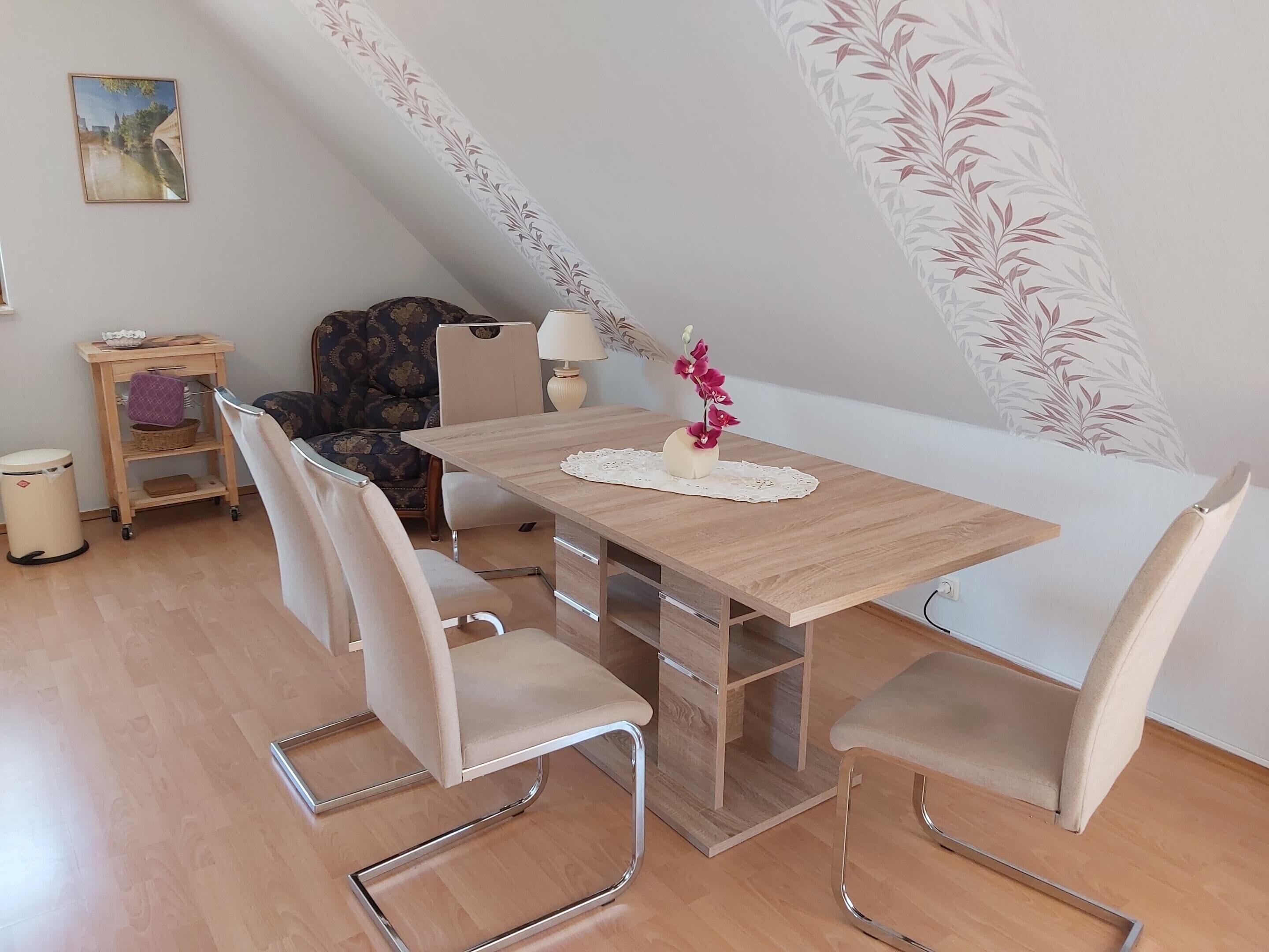 A dining area with a wooden table, four chairs, a vase with flowers, a lamp, and a small shelf with a basket and a bowl.