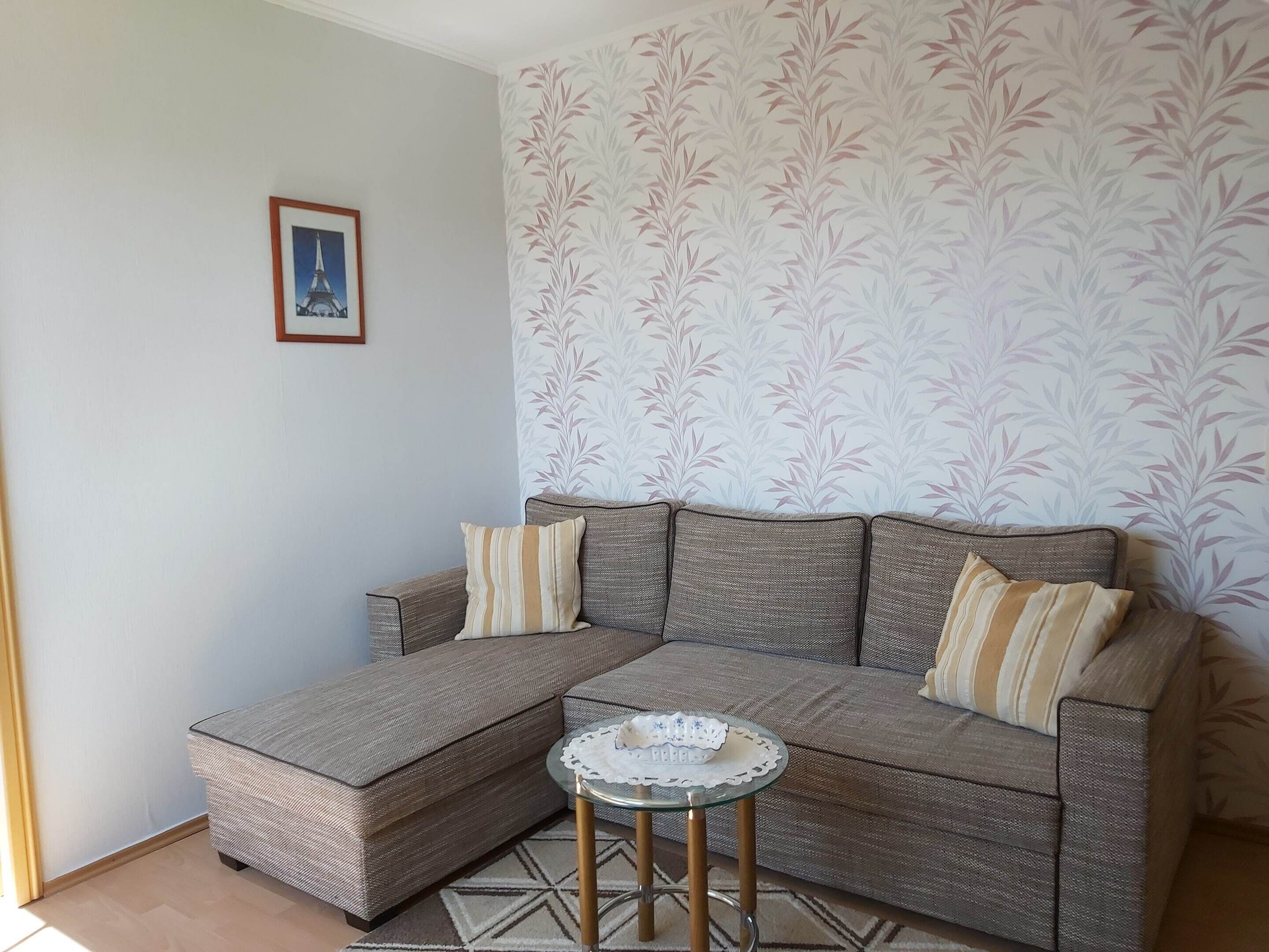 A corner sofa with a glass coffee table, patterned wallpaper, and a framed picture on the wall.