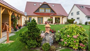 Uma casa de dois andares com telhado de telhas vermelhas, varanda coberta com pilares amarelos e um jardim bem cuidado com diversas plantas e um candeeiro de pedra decorativo.