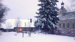 A snow-covered landscape with a building featuring a bell tower and a snow-covered tree. - Holiday Home in Odeigne With Sauna (Manhay)