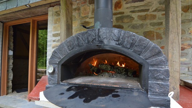 A stone-built outdoor pizza oven with a fire inside, set against a rustic stone wall. - Holiday Home in Odeigne With Sauna (Manhay)